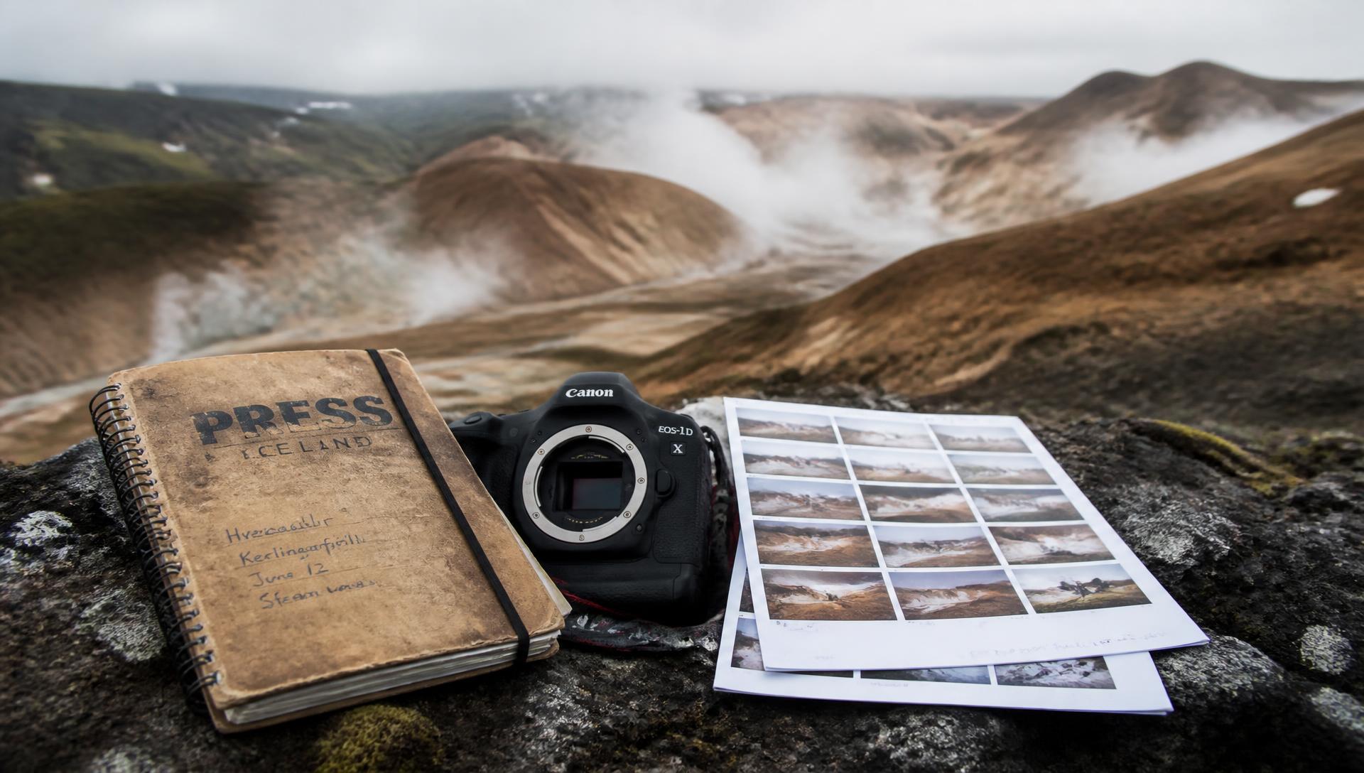 A press field notebook, camera body and printed contact sheets on a basalt ledge above a steaming geothermal valley